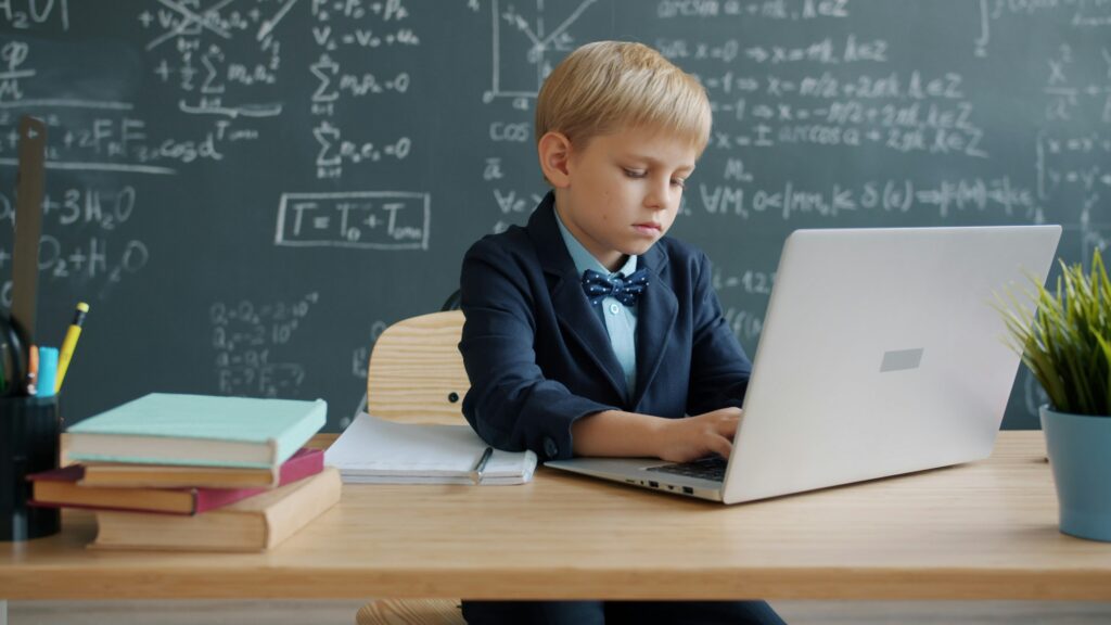 smart little kid using laptop typing then writing at desk in university classroom, blackboard with formulas is visible in background.